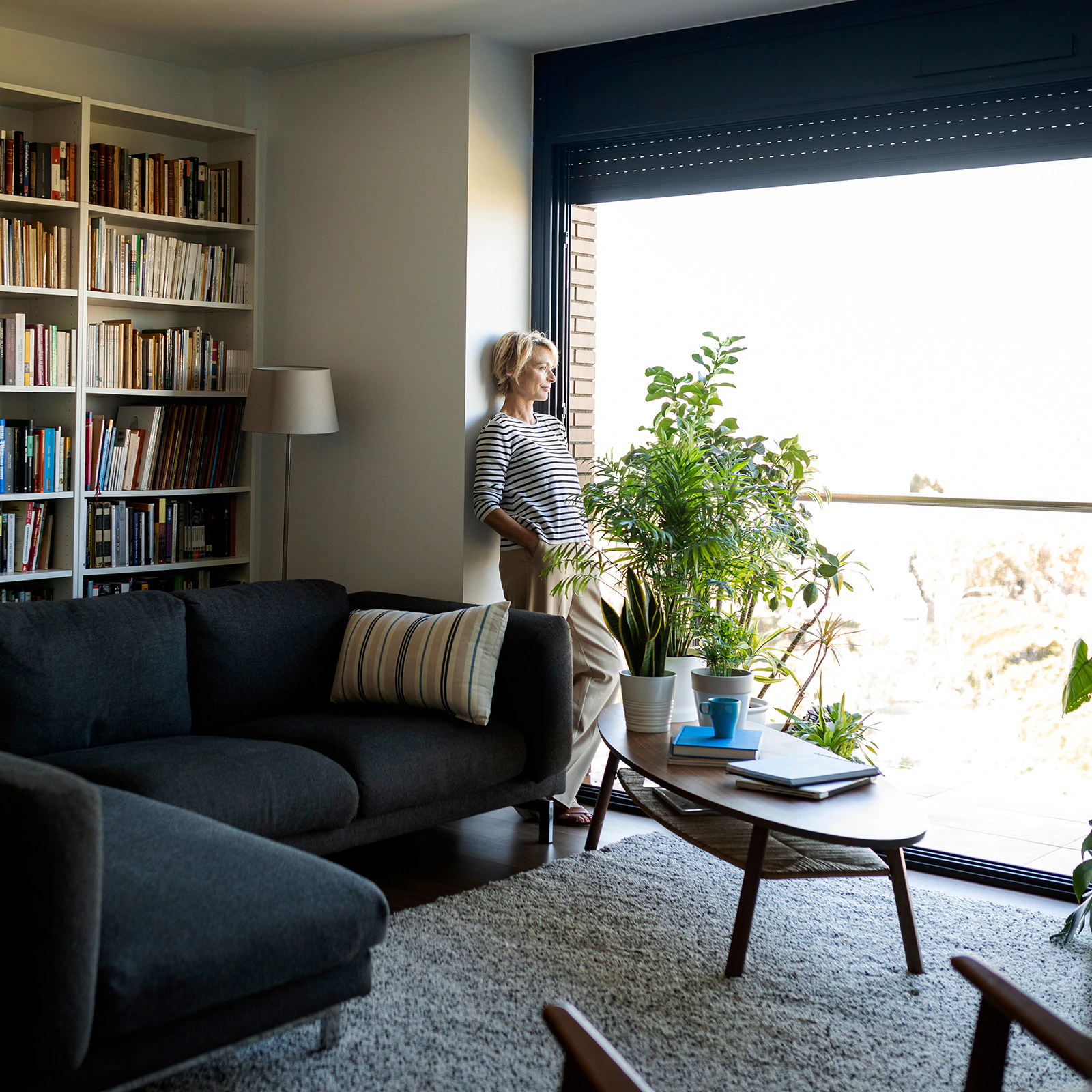 "Woman standing by a large window in a modern living room filled with natural light, surrounded by indoor plants, bookshelves, and cozy furniture.